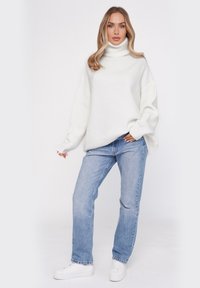 Oversized biały sweter z golfem o prążkowanej fakturze, w połączeniu z jasnoniebieskimi dżinsami o prostym kroju i białymi trampkami.