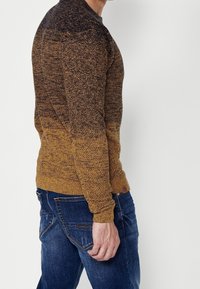 Homme portant un pull moucheté marron et orange foncé et un jean bleu, debout de profil devant un fond clair uni.