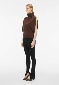 Brūna tīkota turtlenecks ar asimetrisku dizainu, apvienota ar melniem pieguļošiem biksēm ar šķēlumiem apakšā un brūnām papēžu kurpēm.