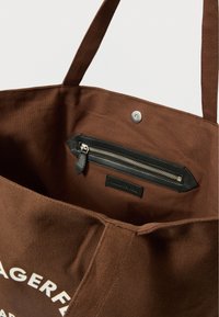 Geantă tote din pânză maro, cu suprafață texturată, curele lungi și un buzunar interior negru cu fermoar și șnur din piele.