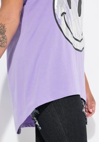 T-shirt en coton violet avec un motif de smiley argenté, doté d'un design d'ourlet incurvé. Associé à un jean en denim noir déchiré.