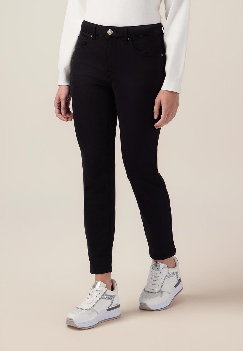 Svarta slim-fit jeans med knappstängning framtill och sidofickor, kombinerat med vita sneakers med silverdetaljer och en kraftig sula.
