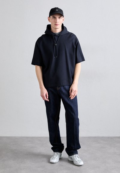 Námornícky oversized mikina s predným vreckom a šnúrkou, kombinovaná s tmavými džínami a bielymi teniskami. Čierna čiapka dokončuje vzhľad.