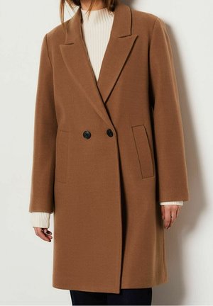 Brun dobbeltradet uldfrakke med sorte knapper båret over en hvid ribbet sweater, vist fra skuldre til midt på låret.