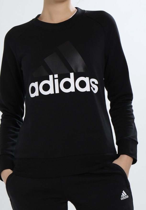 Personne portant un sweat-shirt et un pantalon Adidas noirs avec des logos Adidas blancs, debout les mains dans les poches devant un fond uni.