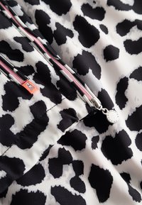 Zwart-wit luipaardprint stof met een zilveren rits met een ronde trekker en een klein oranje label met een hondenlogo.