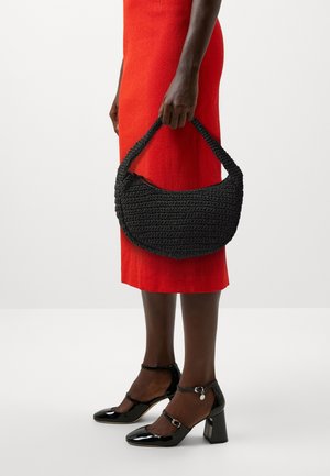 Henkilö, joka pukeutuu punaiseen hameeseen ja mustiin kiiltonahkaisiin korkokenkiin, pitää mustaa kudottua käsilaukkua kahvasta valkoista taustaa vasten.