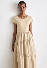 Beige gelaagde maxijurk met een aansluitende top en korte mouwen, voorzien van een ronde hals en een zachte stofstructuur. Subtiele branding op de borst.