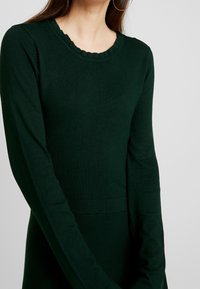 Mörkgrön långärmad tröja i ett mjukt tyg. Har en ribbad rund halsringning med en vågig kant och en åtsittande silhuett.