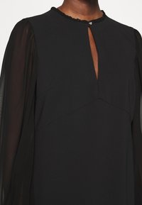 Robe noire avec manches longues transparentes, encolure goutte d'eau fermée par un bouton décoratif unique et légère finition à volants au col.
