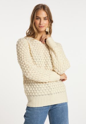 Junge Frau mit langen Haaren, die einen cremefarbenen, strukturierten Strickpullover und blaue Jeans trägt, posiert mit einer Hand nahe am Kinn.