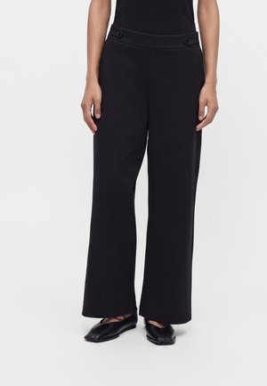 Person wearing black wide-leg pants with button details at the waist and black flat shoes against a plain white background.