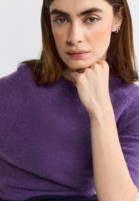 Purple knitted sweater with a textured finish. The model rests her chin on a fist, showcasing a relaxed neckline and side-swept hair.