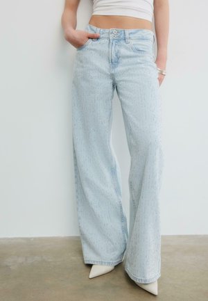 Persona che indossa jeans larghi celeste chiaro con dettagli sottili di strass, scarpe bianche a punta e top corto bianco.