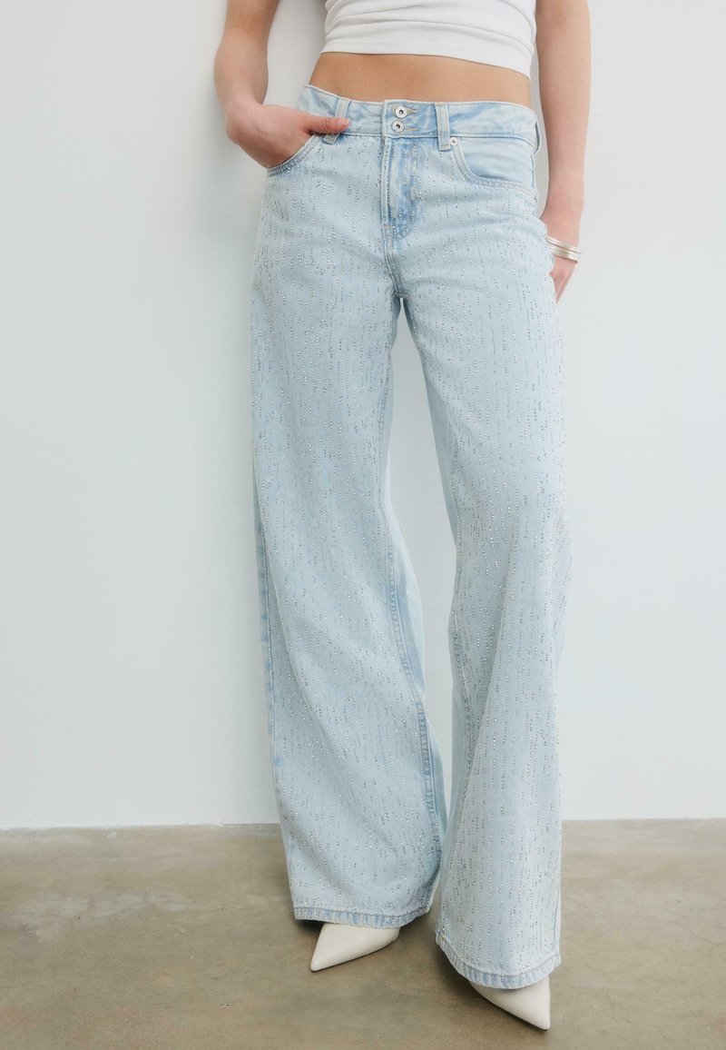 Osoba nosící světle modré džíny s širokými nohavicemi a jemnými detaily z kamínků, bílé špičaté boty a bílý krátký top.
