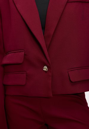 Blazer bordeaux à coupe courte, avec deux poches avant et une fermeture à un bouton. Tissu lisse avec une coupe structurée.