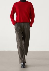 Punainen neulepaita, jossa on pyöreä pääntie, on yhdistetty khaki-värisiin laskostettuihin housuihin. Mustat loafers-kengät täydentävät asukokonaisuuden. Pintamateriaalit ovat sileitä.