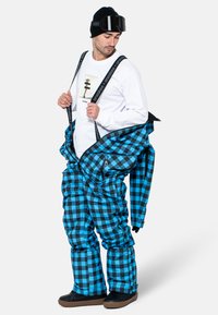 Pantalones de esquí a cuadros azules y negros con tirantes ajustables, bolsillos con cremallera y corte holgado, usados sobre una camiseta blanca de manga larga y un gorro negro.