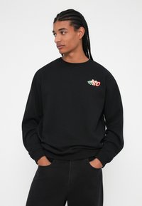 Schwarzer Sweatshirt mit runder Halslinie und langen Ärmeln, versehen mit einem farbenfrohen gestickten Design auf der linken Brust. Lässige Passform mit gerippten Bündchen.