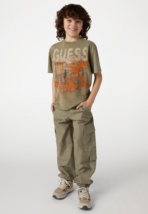 Groene t-shirt met "GUESS" logo en oranje bloemenontwerp, gecombineerd met kaki cargobroek en beige sneakers met witte accenten.