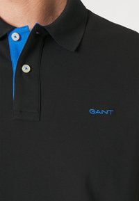 Fekete galléros póló kék gallérral, kétgombos sliccel és a mellrészen kék "GANT" logóval hímzve. Texturált anyag.