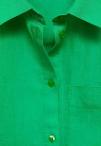 Cămașă verde din in cu guler ascuțit, patru nasturi și un buzunar pe piept. Textură netedă cu detalii de cusătură subtile.