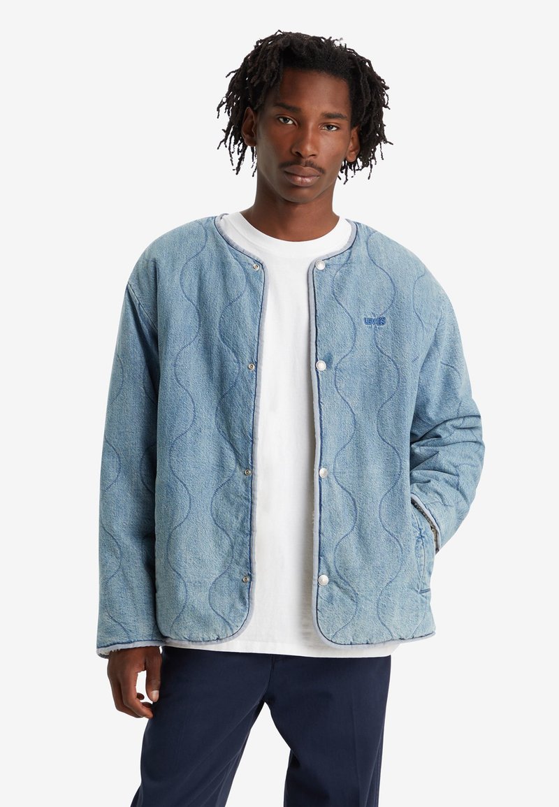 Chaqueta de denim azul claro con un patrón de ondas acolchado, cierre de botones a presión y cuello acanalado, llevada sobre una camiseta blanca.