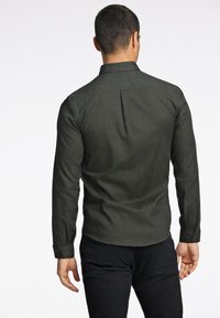 Camisa de manga larga verde oscuro con un acabado texturizado, puños abotonados y un pliegue en la parte posterior, llevada con pantalones negros, mostrada desde la parte de atrás.