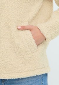 Kurtka polarowa w kremowym kolorze o fakturowanej powierzchni, z boczną kieszenią i miękkim, przytulnym wykończeniem. Rękawy mają lekko postrzępione brzegi.