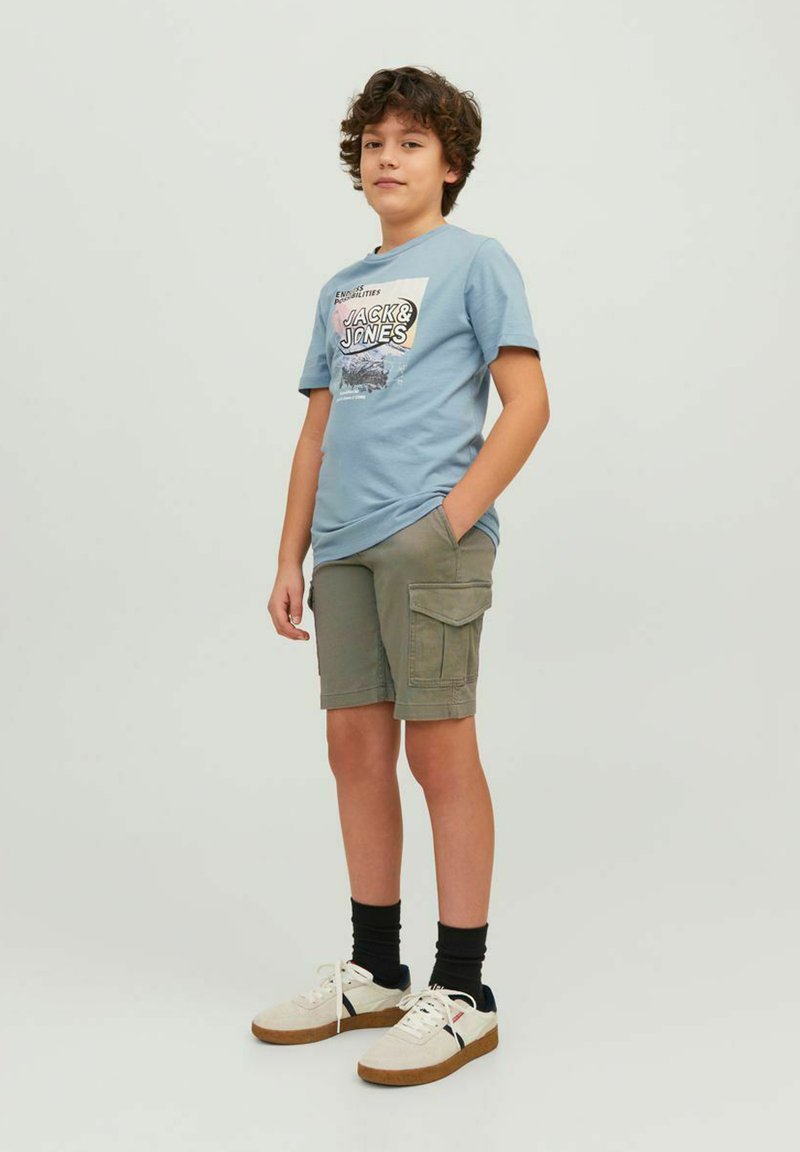 T-shirt in cotone azzurro chiaro con stampa grafica, abbinato a pantaloni cargo verde oliva. Sneakers bianche con suola marrone e calze nere a mezz'asta.