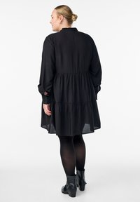 Must mustriga mustmärkidega must kleit, mis on valmistatud kergest kangast, pikkade varrukatega, volangkraega ja nööpidega mansettidega. Kantud opaksete mustade sukkpükste peale.