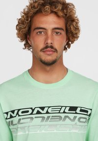 Camiseta de manga corta verde menta con un diseño gráfico en negro y plata que presenta letras alternas en el pecho. Textura suave.