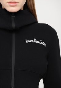 Sweatshirt noir à fermeture éclair avec un col montant, une texture côtelée, et un logo "Versace Jeans Couture" brodé en blanc sur la poitrine.