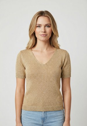 Giovane donna con capelli biondi che indossa un maglione dorato a maniche corte con scollo a V e jeans azzurri, in piedi davanti a uno sfondo chiaro e uniforme.