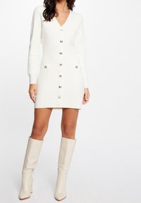 Abito lavorato a maglia bianco con scollo a V, maniche lunghe e bottoni frontali. Presenta due tasche con dettagli metallici. Indossato con stivali beigi al ginocchio.