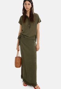 Oliwkowy top z kołnierzykiem i guzikami, w połączeniu z pasującą maxi spódnicą z frędzlami na dolnej krawędzi. Modelka trzyma plecioną torbę.