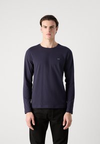 Ναυτικό μακρυμάνικο πουκάμισο από μαλακό ύφασμα, με στρογγυλή λαιμόκοψη και μικρό λογότυπο της μάρκας στο αριστερό στήθος, σε συνδυασμό με μαύρο παντελόνι.