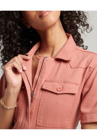 Różowy kombinezon z denimu z krótkimi rękawami, zamkiem z przodu i kieszenią na piersi. Miękka faktura i casualowy design.