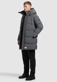 Muškarac u sivoj zimskoj jakni s kapuljačom, crnim hlačama i crnim čizmama stoji protiv jednostavne pozadine.