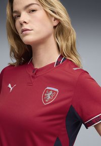 Femme blonde portant un maillot de sport rouge avec des logos Puma blancs et un blason de lion argenté, debout devant un fond gris.