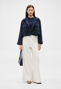 Tânără purtând o jachetă scurtă bleumarin, pulover închis la culoare, pantaloni albi largi, pantofi bej, ținând o geantă bleumarin, stând într-un cadru simplu, cu fundal alb.