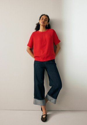 Red short-sleeve top paired with wide-leg dark denim jeans featuring rolled cuffs. The outfit is complemented by black flat shoes.