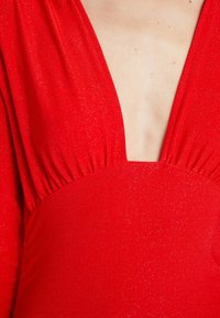 Robe en tissu rouge scintillant avec un profond décolleté en V dévoilant le haut de la poitrine et une couture froncée sous la poitrine.