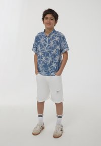 Camicia blu a maniche corte con bottoni, decorata con un motivo di palme bianche, abbinata a pantaloni corti di jeans bianchi con bordo sfrangiato. Indossata con scarpe sportive e calzini.