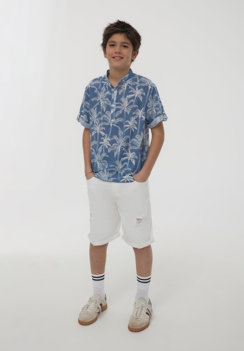 Camicia blu a maniche corte con bottoni, decorata con un motivo di palme bianche, abbinata a pantaloni corti di jeans bianchi con bordo sfrangiato. Indossata con scarpe sportive e calzini.