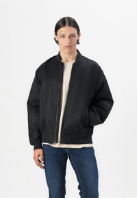 Chaqueta bomber negra con cierre frontal, puños y dobladillo acanalados. Lleva puesta una camisa beige, combinada con jeans de mezclilla azules.