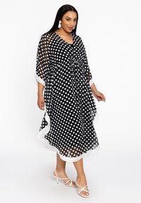 Schwarzes kimono-artiges Kleid mit Punkten, einer Taillenschnürung, transparentem Material, weißem Fransenbesatz und weiten Ärmeln, kombiniert mit weißen Straps-Heels.
