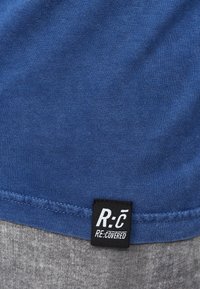 Sininen T-paita, jolla on sileä pinta, ja alasaumassa musta etiketti, jossa valkoiset kirjaimet "R:C RE:COVERED".