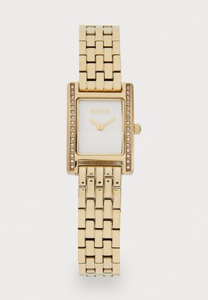 Montre rectangulaire dorée avec un cadran blanc, présentée avec une lunette ornée de cristaux et un bracelet à maillons en métal doré.