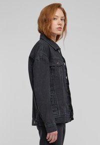 Vöröshajú személy, aki fekete farmerdzsekit visel, oldalnézetben áll sima világosszürke háttér előtt.